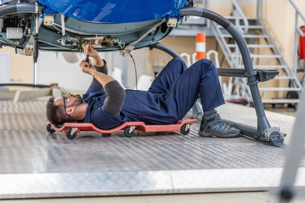 Aviation engineer lying on cart on wheels and fixing helicopter engine with work tool, wide shot